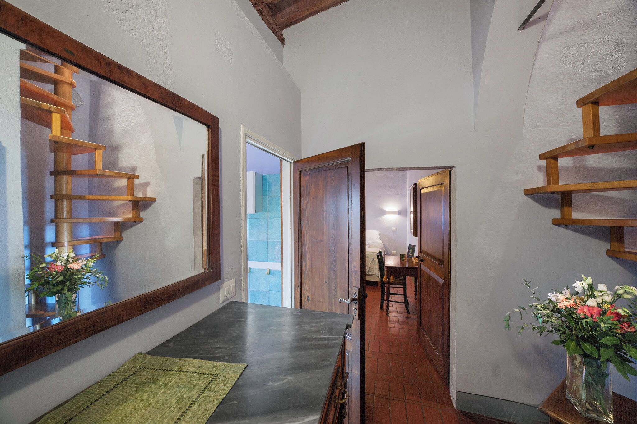 A narrow hallway with a wooden staircase, a dining area with a table and chairs, and a vase with flowers.