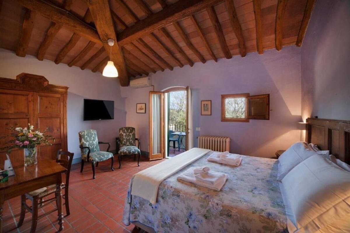 A bedroom with a wooden ceiling, a bed with a floral bedspread, a wooden desk with a vase of flowers, two chairs, and a window with a view of the outdoors.
