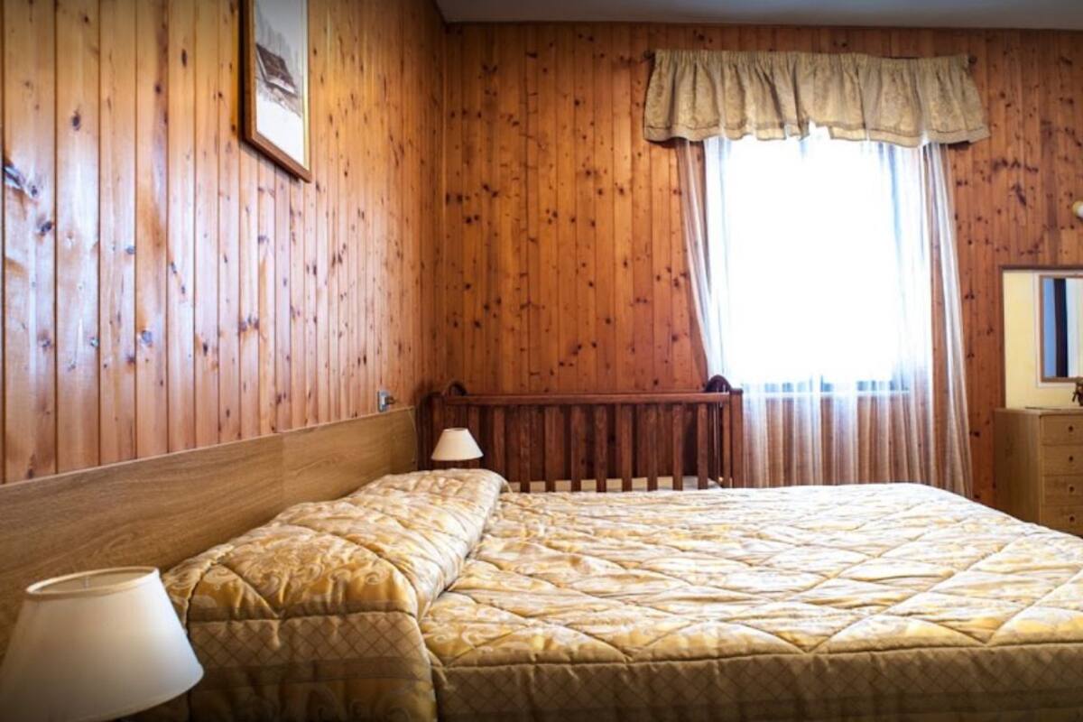 A room with wooden paneling, a bed with a quilt, a nightstand with a lamp, a dresser, and a window with curtains.