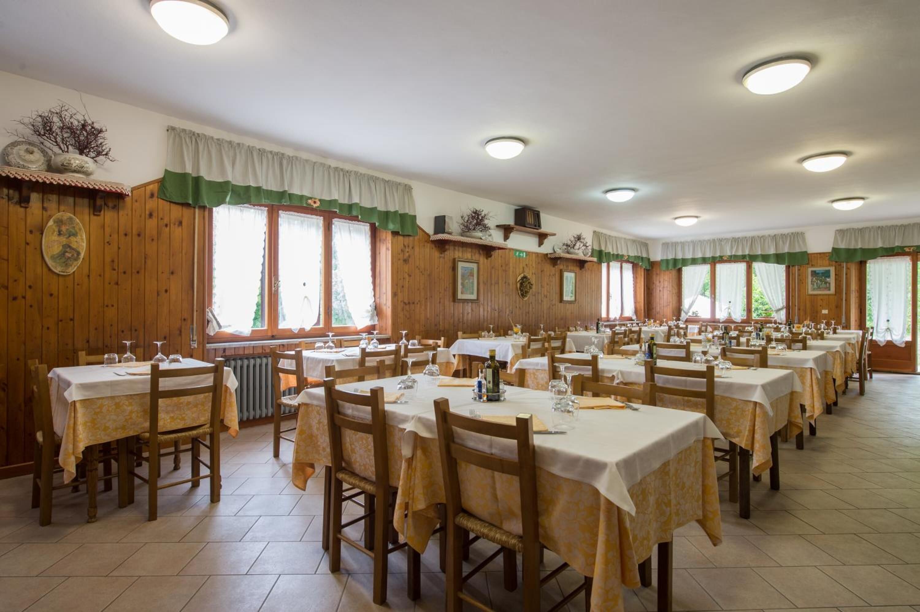 A large dining hall with multiple tables and chairs, wooden walls, and large windows with curtains.