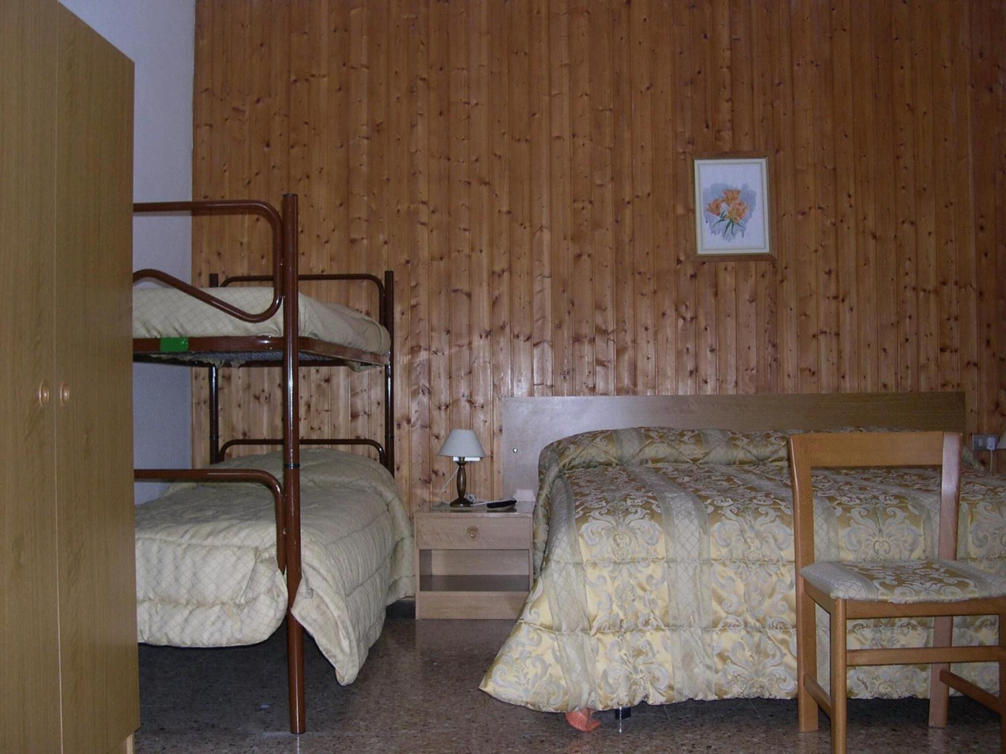 Una stanza con letti a castello, un letto singolo, una sedia, un comodino con una lampada e un quadro appeso al muro.