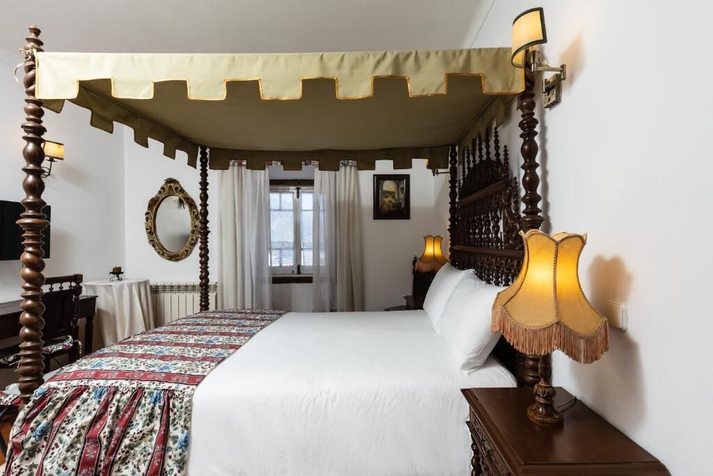 A four-poster bed with a canopy, a bedside lamp, a mirror, and a window with curtains.