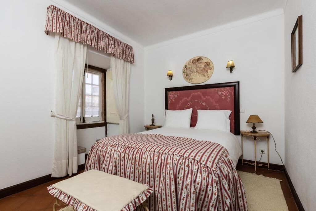 A bedroom with a bed, a window with curtains, a bedside table with a lamp, and a wall-mounted artwork.