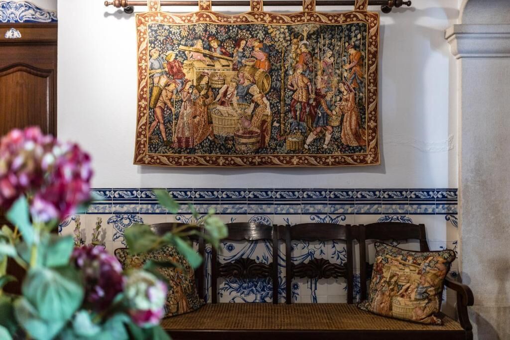 A room with a large tapestry, a wooden bench with a cushion, and a tiled wall with a blue and white pattern.