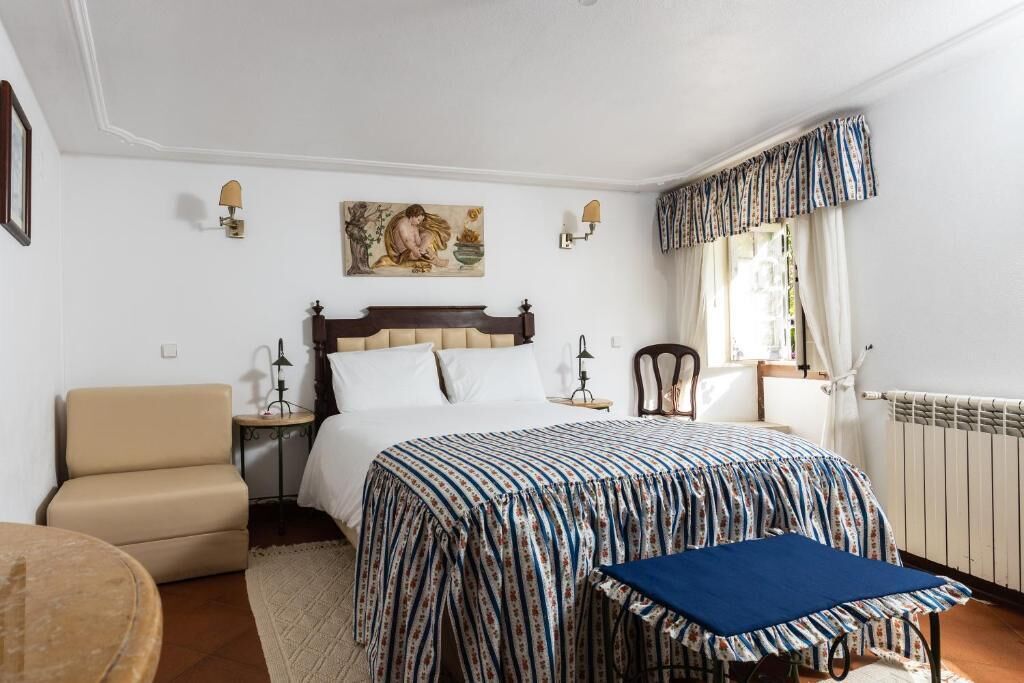 A bedroom with a bed, a chair, a table, and a window with curtains.