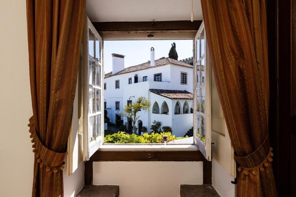 Vista a través de una ventana abierta con cortinas, mostrando un edificio con paredes blancas y tejido de tejas.