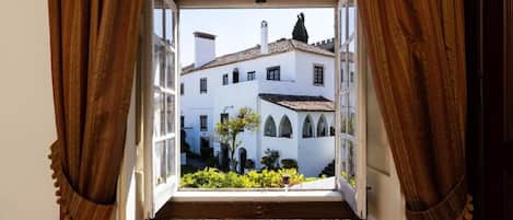 Vista a través de una ventana abierta con cortinas, mostrando un edificio con paredes blancas y tejido de tejas.