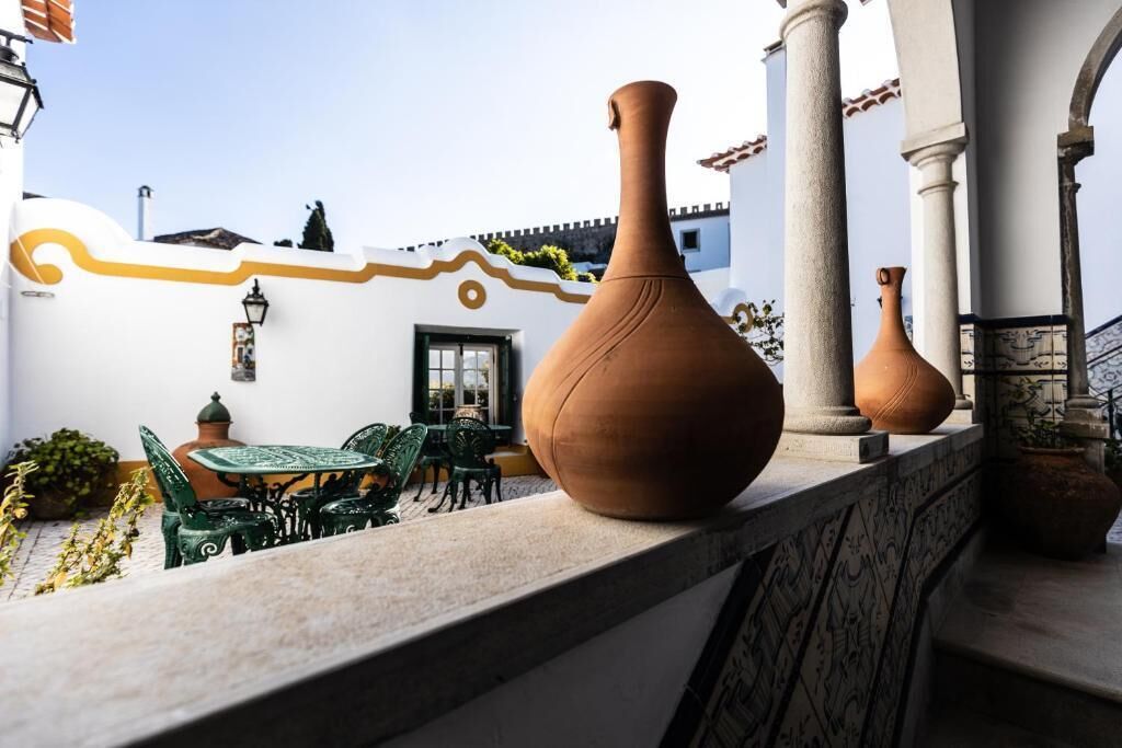 Terraço com vasos de terracota, mesa com cadeiras e um edifício branco com uma linha amarela decorativa.
