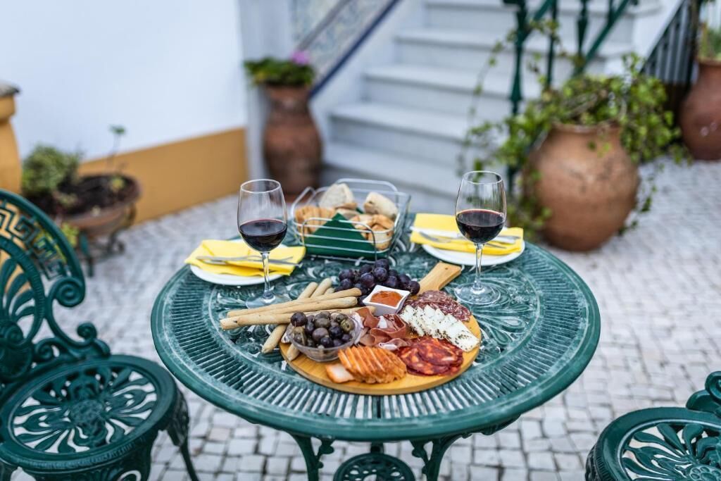 Ein runder Tisch mit einer Wurstplatte, zwei Weingläsern und grünen Gartenmöbeln.