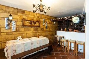 Una habitación rústica con pared de piedra, un banco de madera, una mesa con mantel floral y una zona de bar con taburetes.
