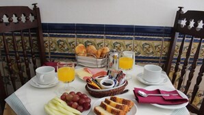 A breakfast table with plates of fruit, bread, and juice, set in a room with patterned tile walls and wooden chairs. - Casa de S. Thiago do Castelo (Obidos)