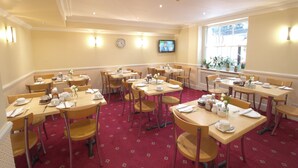 A hotel dining area with multiple tables set for breakfast, equipped with chairs, and a television mounted on the wall. - Balmoral House Hotel (London)