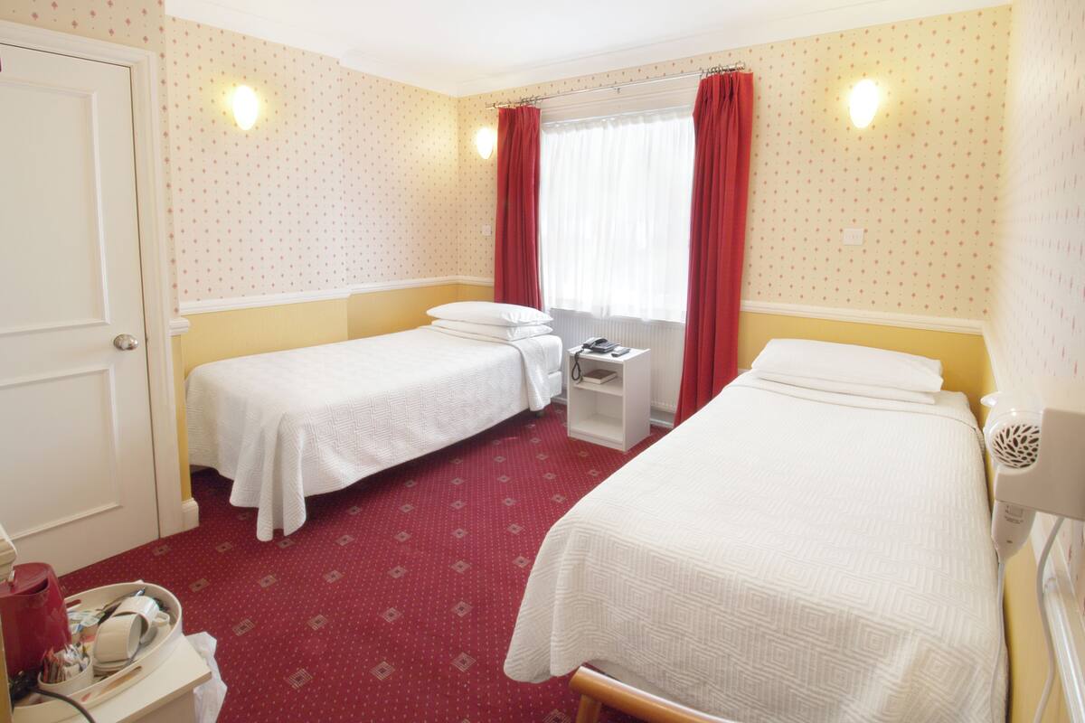 A hotel room with two single beds, a small desk with a lamp, and a window with curtains.