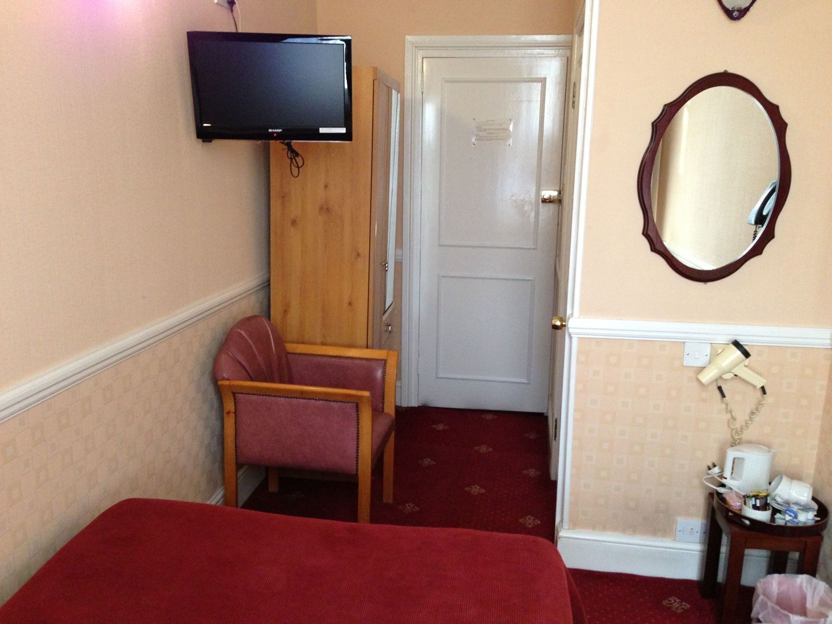 A hotel room with a bed, a television mounted on the wall, a chair, a door, a mirror, and a small table with a hairdryer and other items.