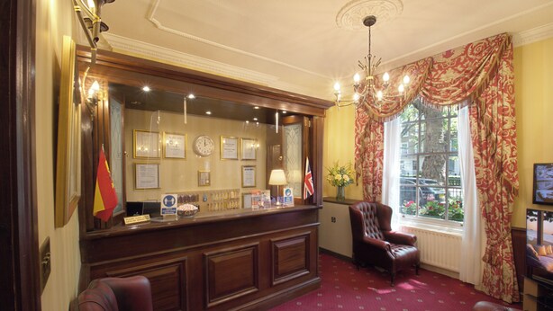 A reception area with a wooden desk, framed certificates, and a flag. There are leather chairs and a window with curtains. - Balmoral House Hotel (London)