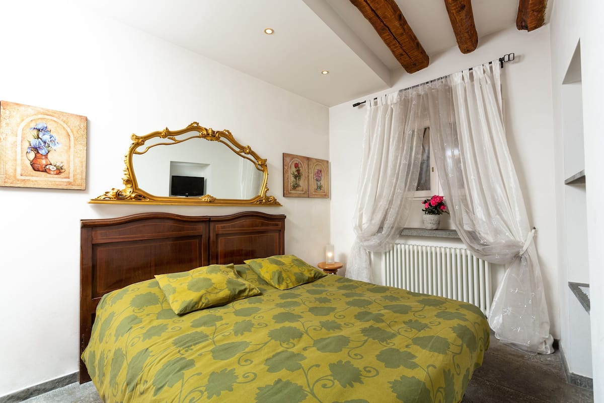 A bedroom with a bed, a wooden headboard, a mirror, a window with sheer curtains, and a radiator.