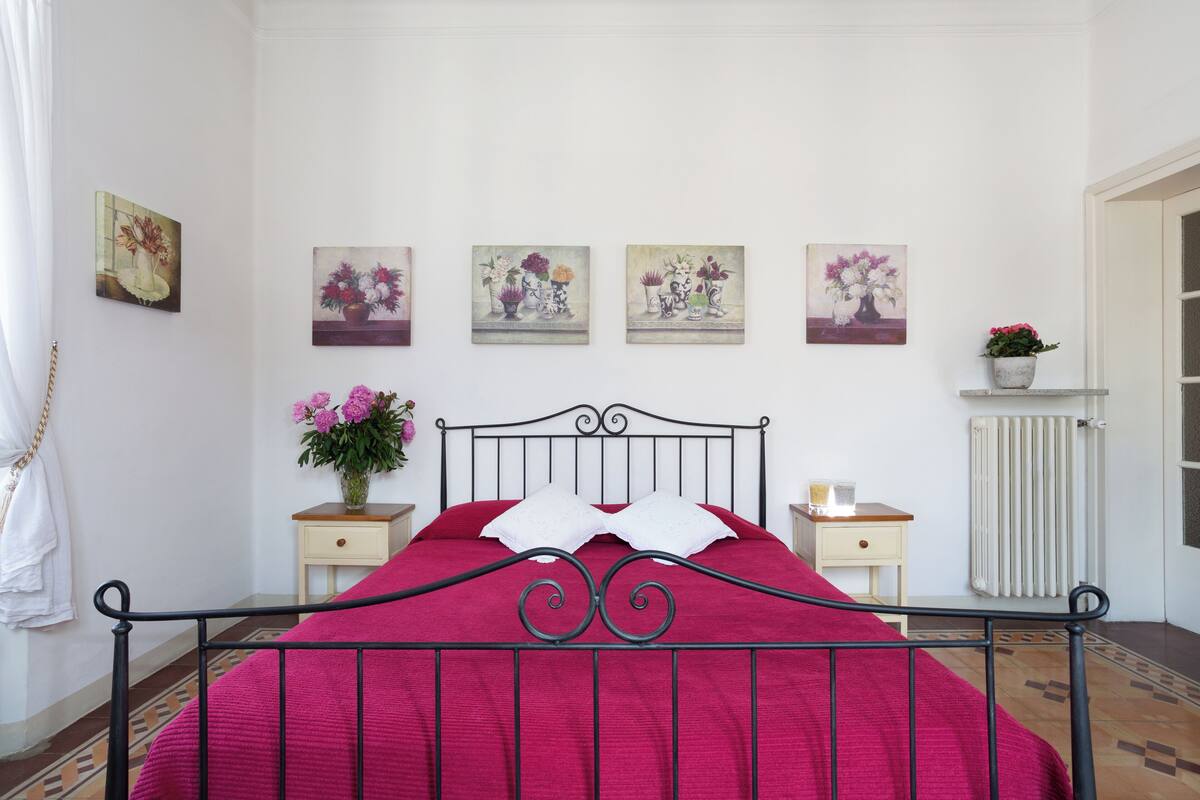 A neatly arranged bedroom with a metal bed frame, a red bedspread, and a white pillow. There are four paintings on the wall, each depicting floral arrangements. A small potted plant is on a nightstand beside the bed.
