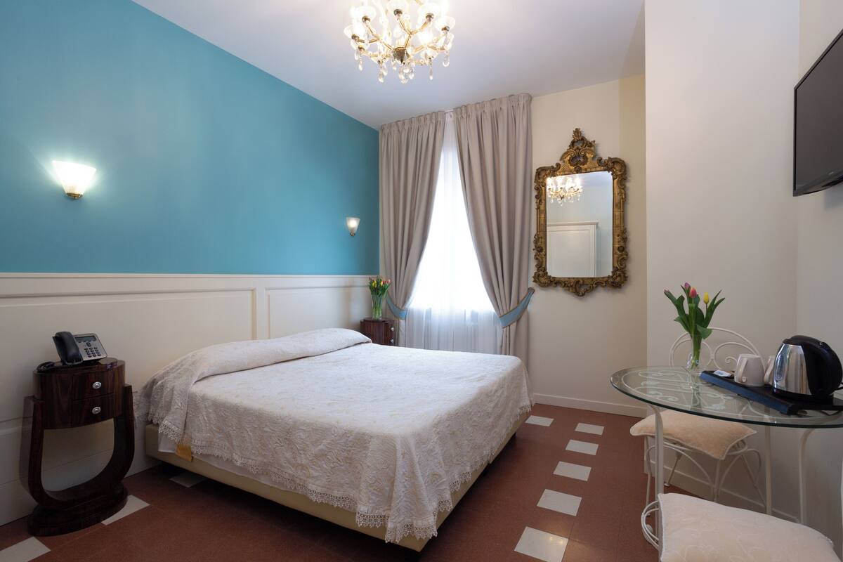 A hotel room with a bed, a bedside table with a phone, a glass table with a kettle, and a mirror on the wall.
