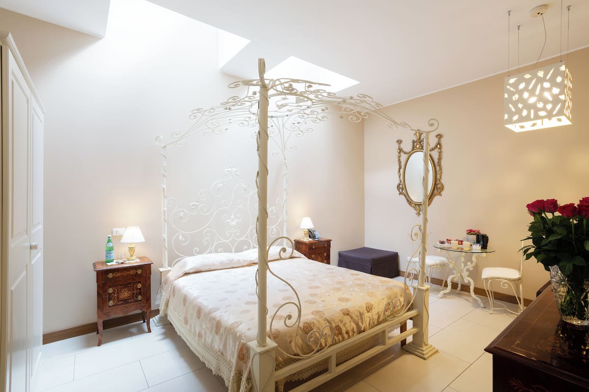 A four-poster bed with a canopy, a bedside table with a lamp, a small round table with a chair, and a vase with flowers.