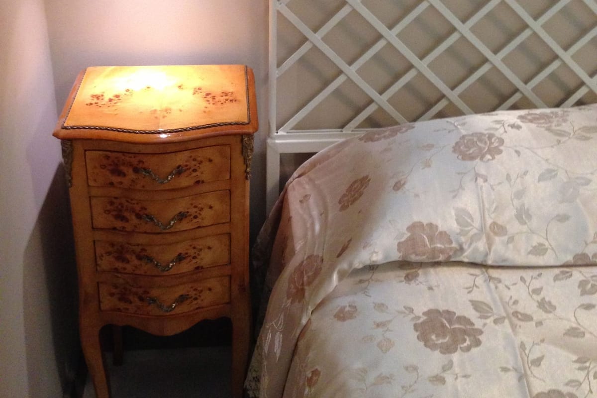 A bedroom with a wooden nightstand, a bed with a floral bedspread, a white headboard, and a wall-mounted light.