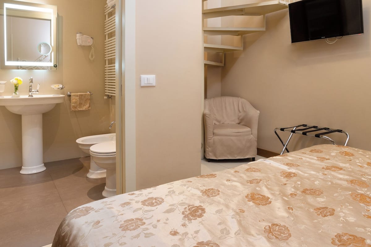 A bedroom with a bed, a TV mounted on the wall, a chair, and a bathroom visible through an open door.