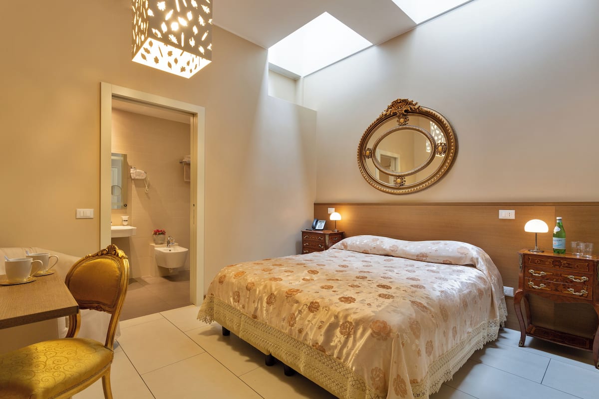 A bedroom with a bed, a bedside table, a mirror, a chair, a dresser, a lamp, and a skylight.