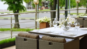 An outdoor dining area with wicker furniture, white tablecloths, and a view of a tree-lined street.