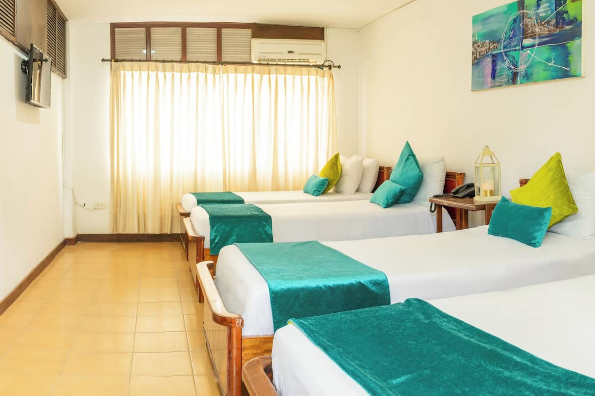 A hotel room with four single beds, each with a teal bedspread and pillows, a wooden headboard, a bedside table with a lamp, and a window with curtains.
