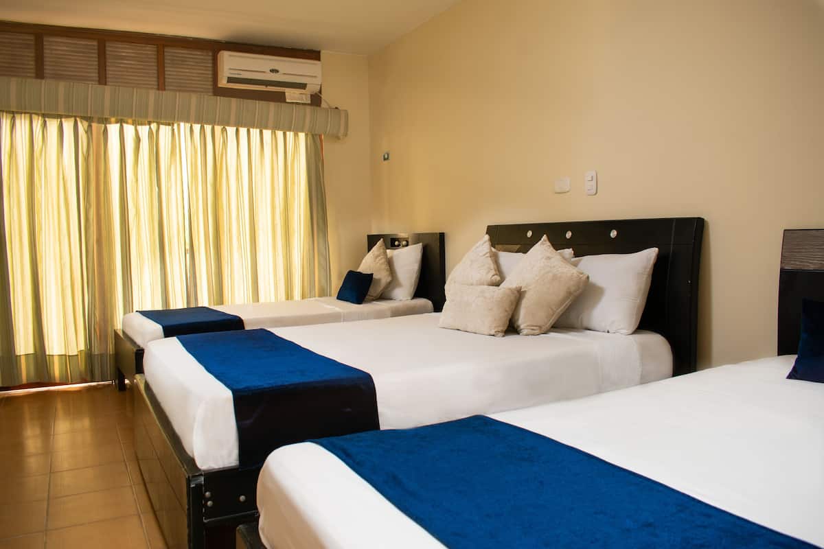 A hotel room with three beds, each with a blue bedspread and white linens. There is an air conditioning unit on the wall and a window with curtains.