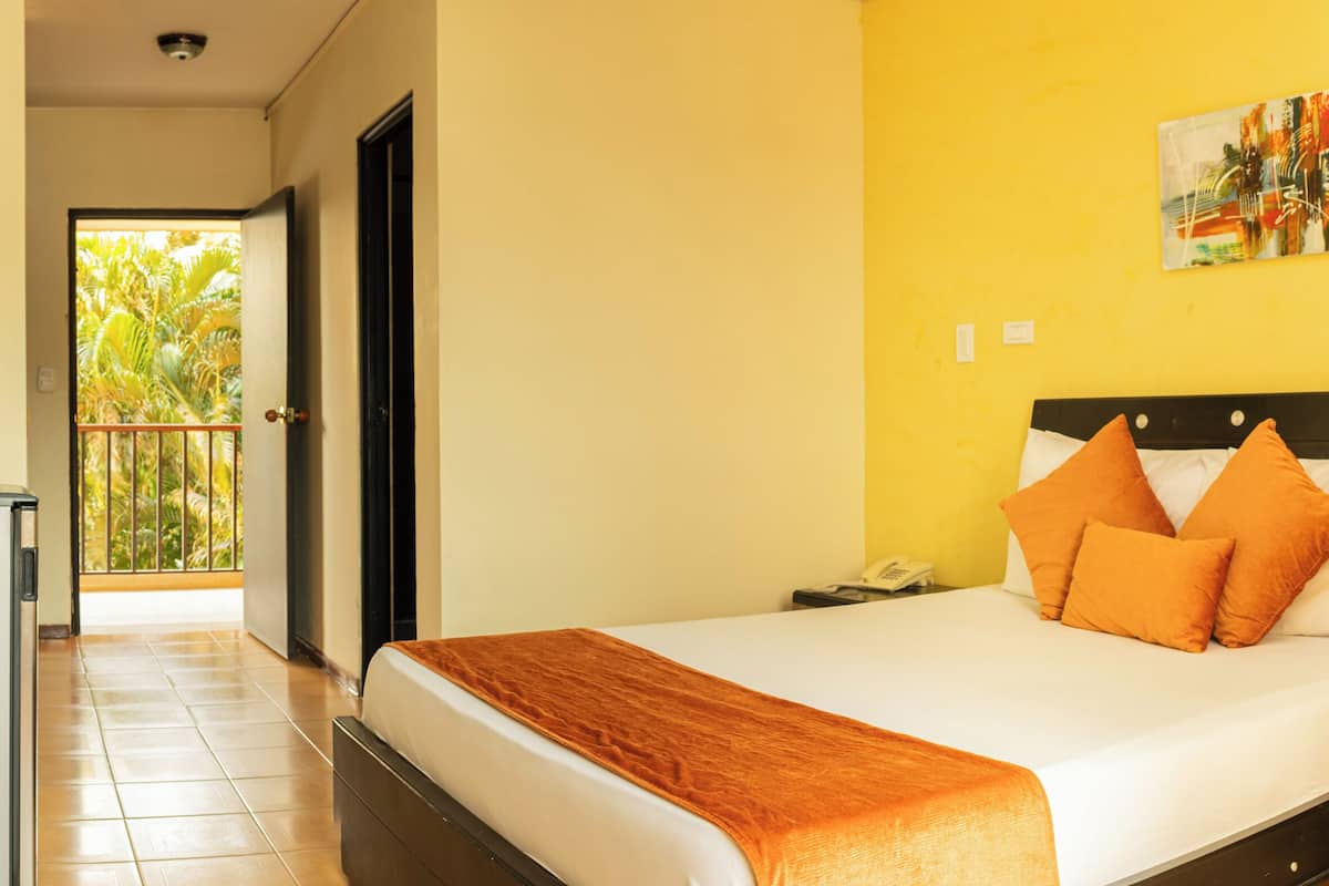 A hotel room with a bed, a desk, a chair, a small refrigerator, and a balcony with a view of greenery.