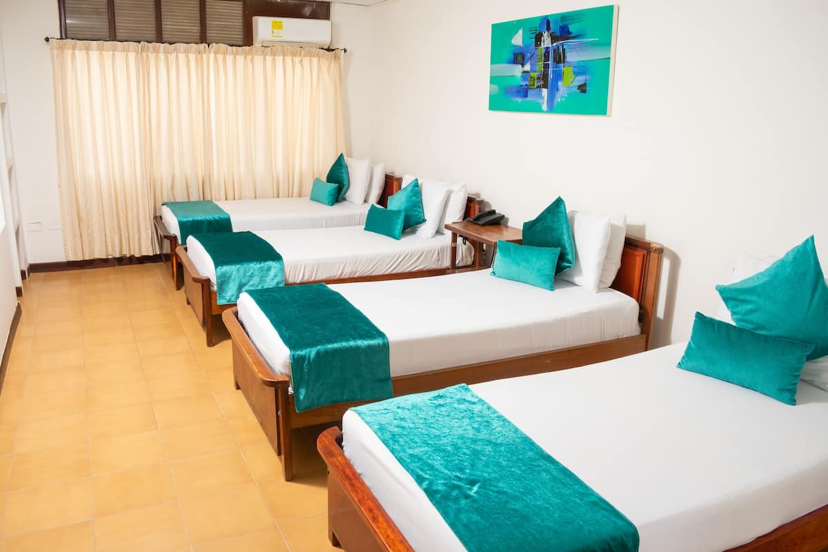 A hotel room with four single beds, each with a teal bedspread and pillows, a wooden nightstand, a desk with a chair, and a window with curtains.