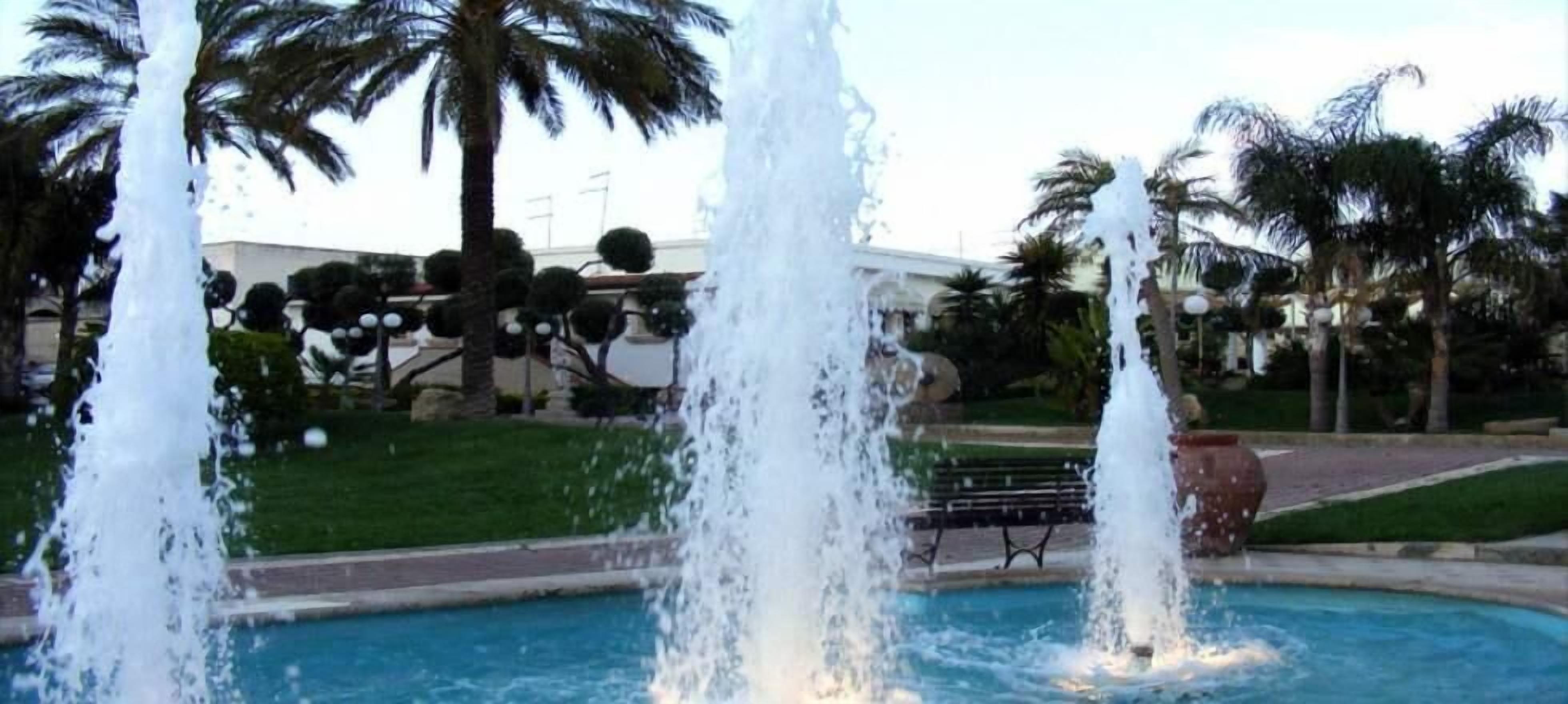 Une fontaine dotée de plusieurs jets d’eau, située dans un espace piscine, entourée de palmiers et avec un bâtiment en arrière-plan.