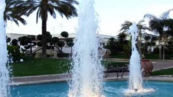 Une fontaine dotée de plusieurs jets d’eau, située dans un espace piscine, entourée de palmiers et avec un bâtiment en arrière-plan.