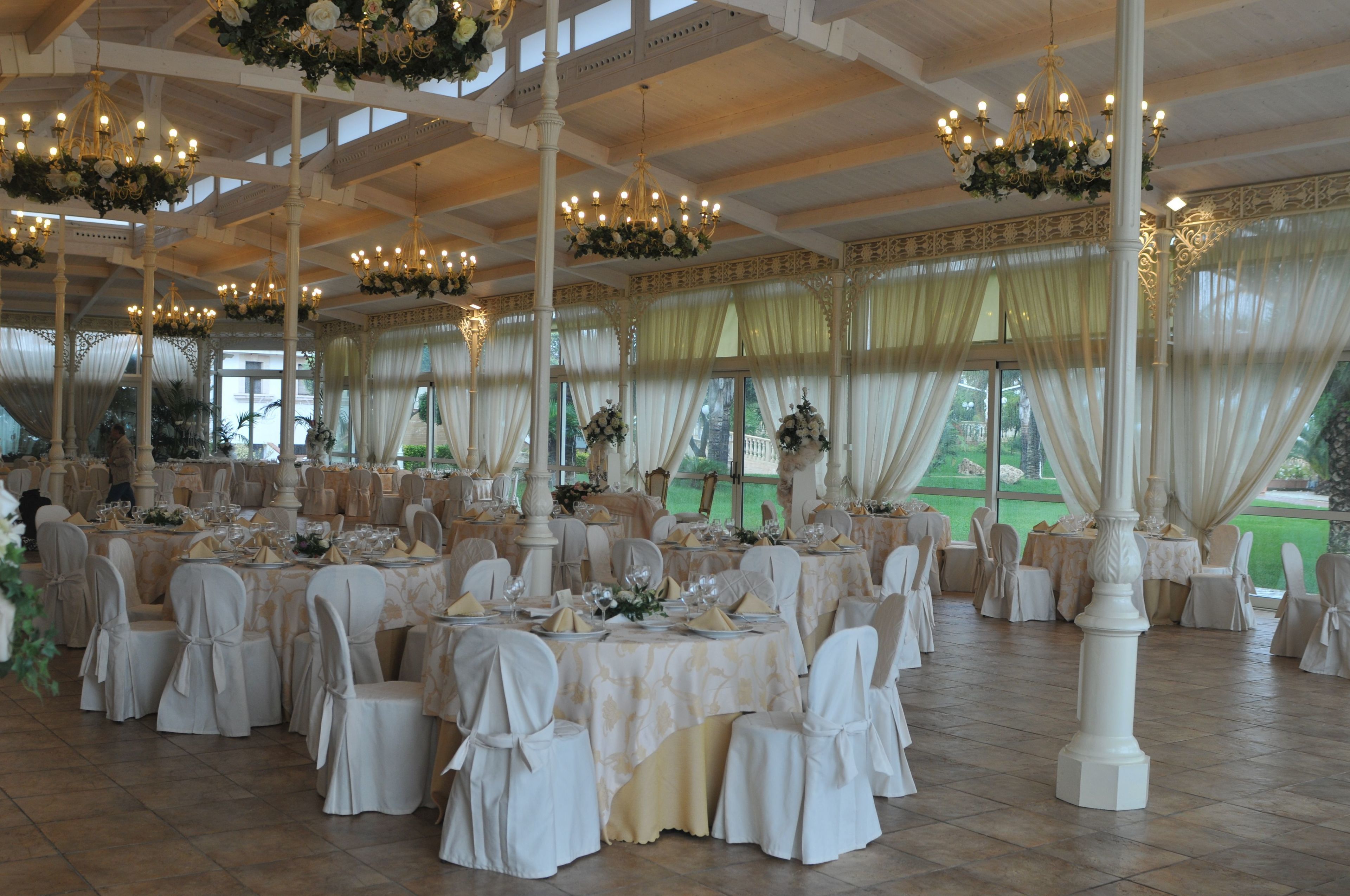 Un salón de banquetes con mesas redondas, sillas blancas, candelabros y ventanas con cortinas.