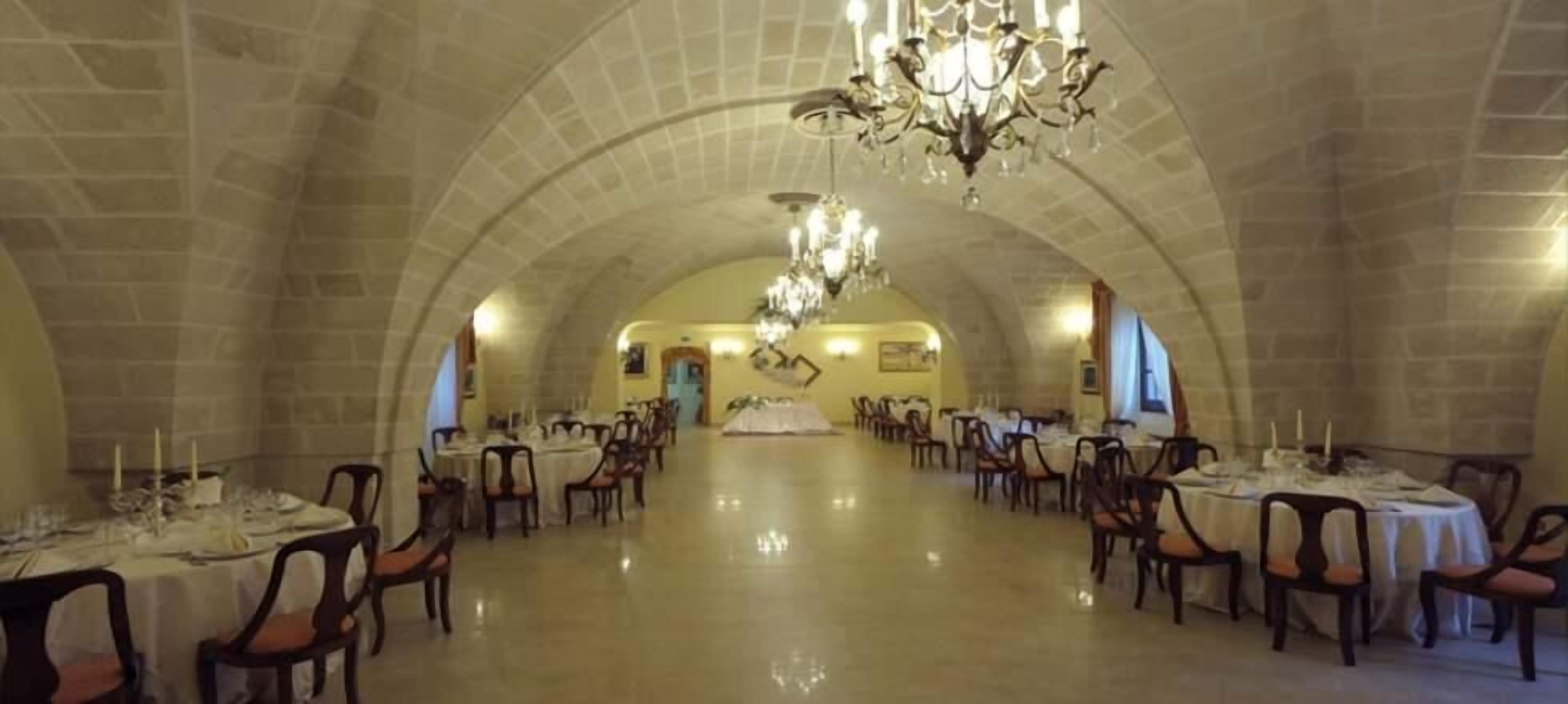Une vaste salle à manger dotée de hauts plafonds en arc de cercle et de lustres. Les tables sont dressées avec des nappes blanches et des bougies.