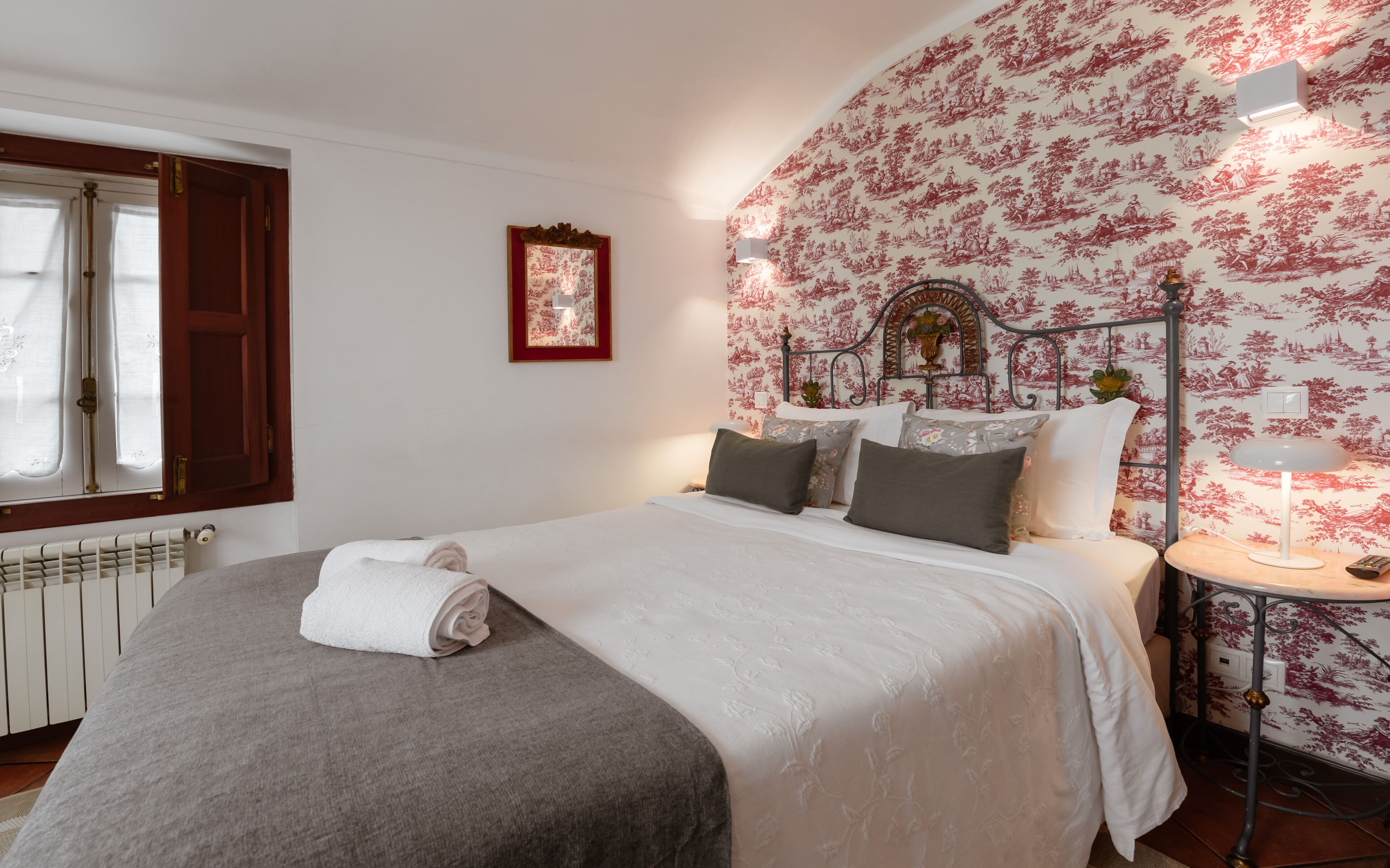 Un lit bien fait, avec une serviette roulée, un radiateur, un mur recouvert d’un papier peint à motifs floraux et une table de chevet avec une lampe.