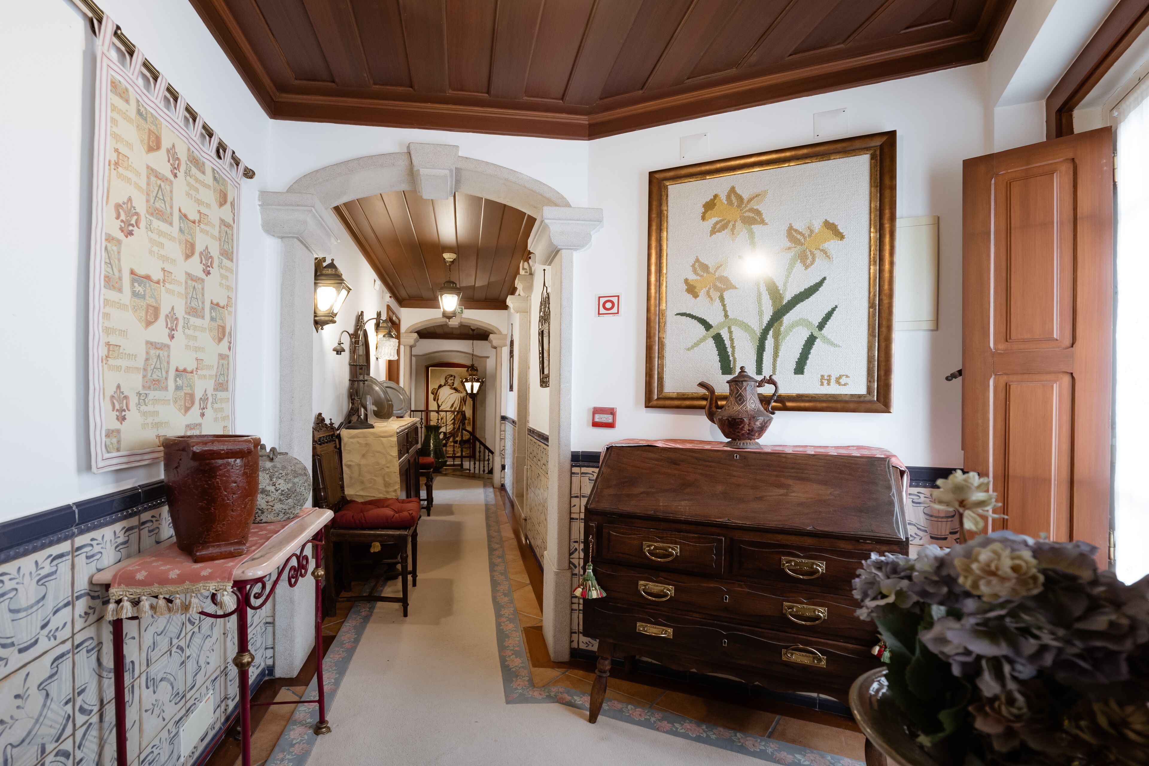 Un pasillo con piso de baldosas, un aparador de madera, un cuadro grande con motivos florales y una puerta roja.