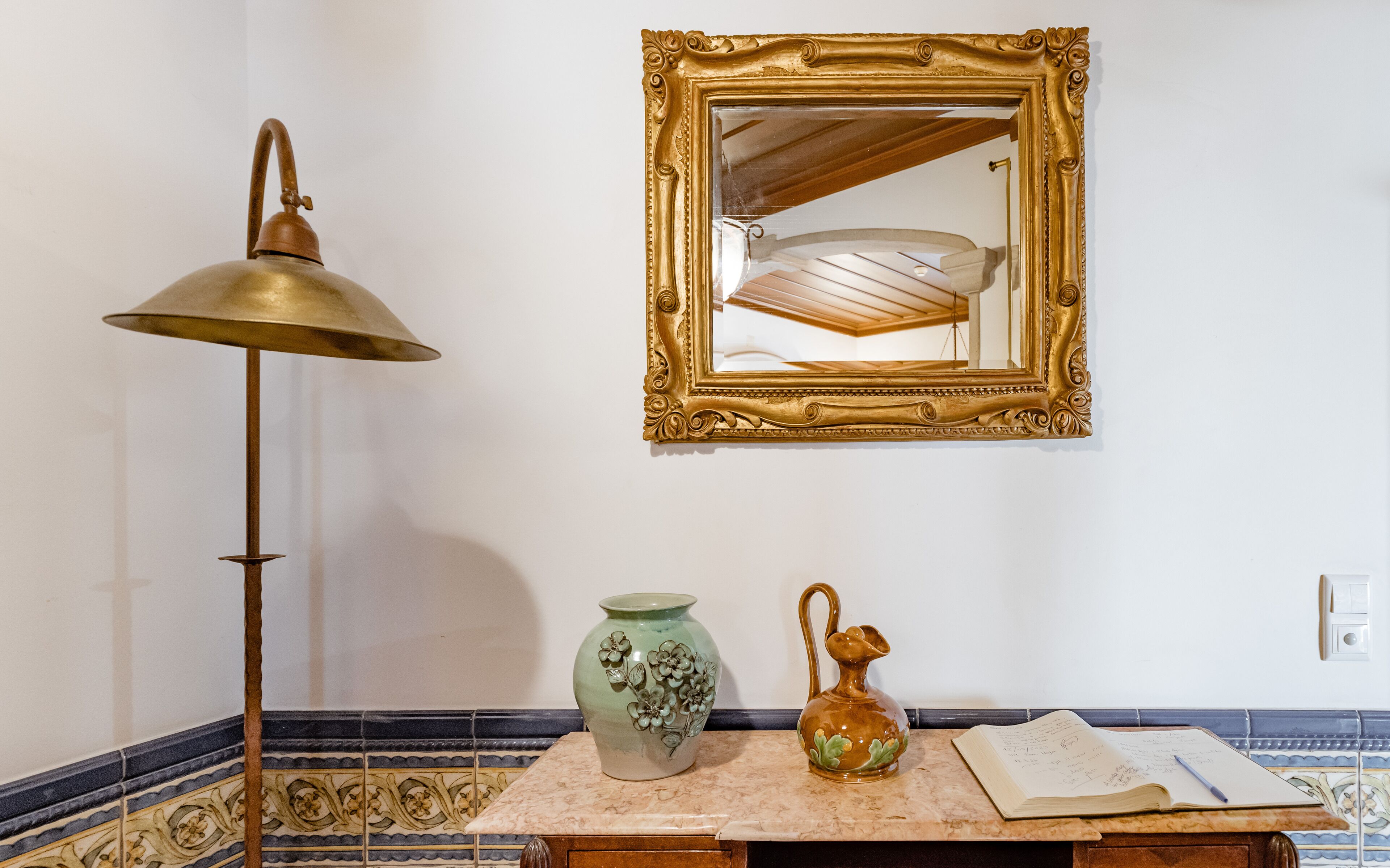 A room with a lamp, a mirror, a vase, a pitcher, and a notebook on a tiled countertop.