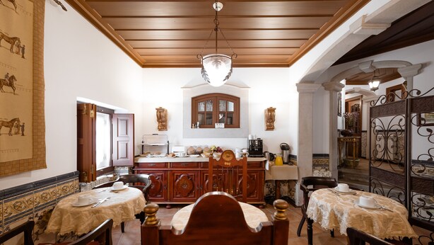A traditional dining room with wooden furniture, a chandelier, and a kitchen area with a window above the counter. - Casa de Sao Thiago d' Obidos (Obidos)