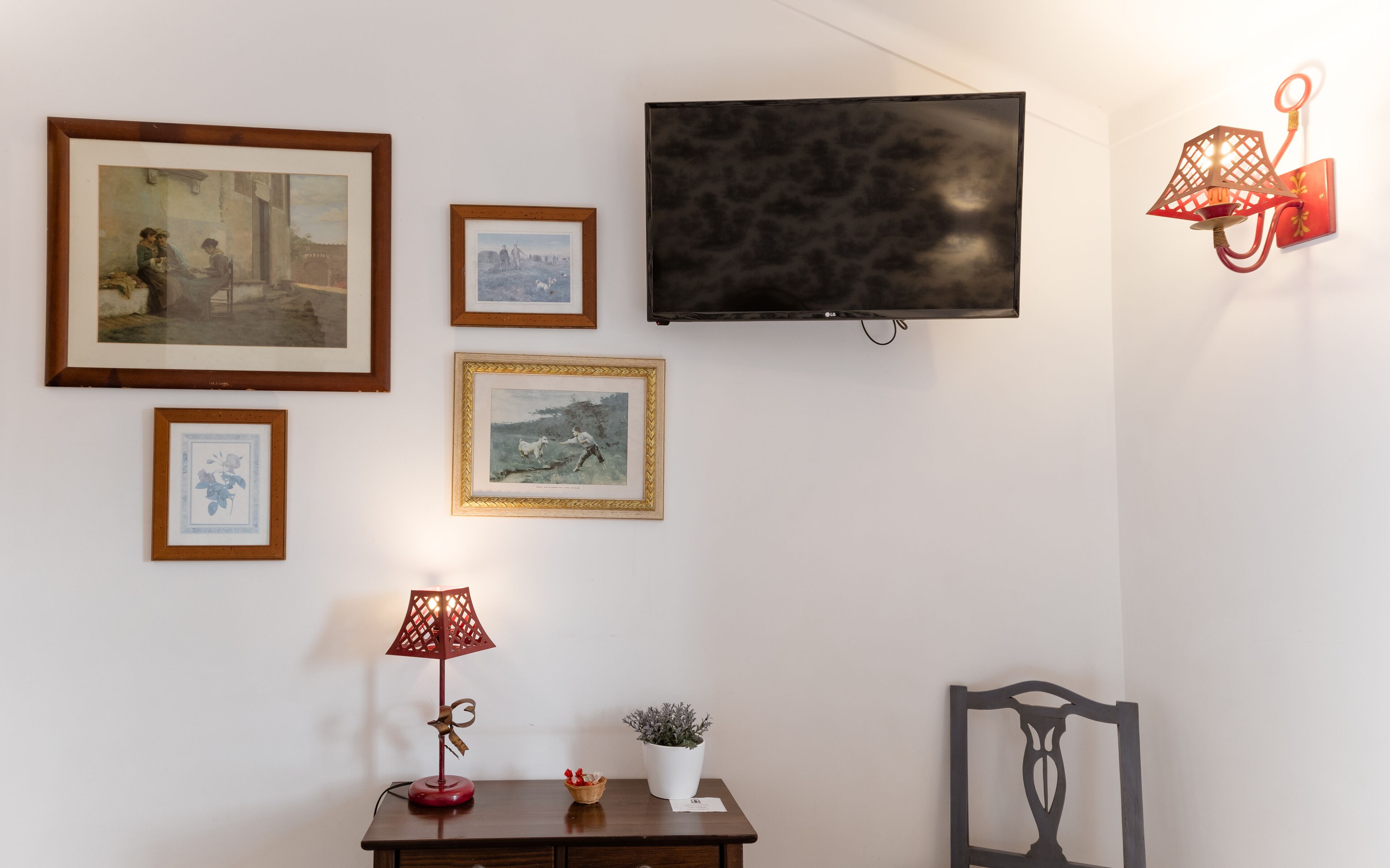 A room with a flat-screen TV mounted on the wall, a wooden cabinet with a small plant, a red lamp, and several framed pictures.