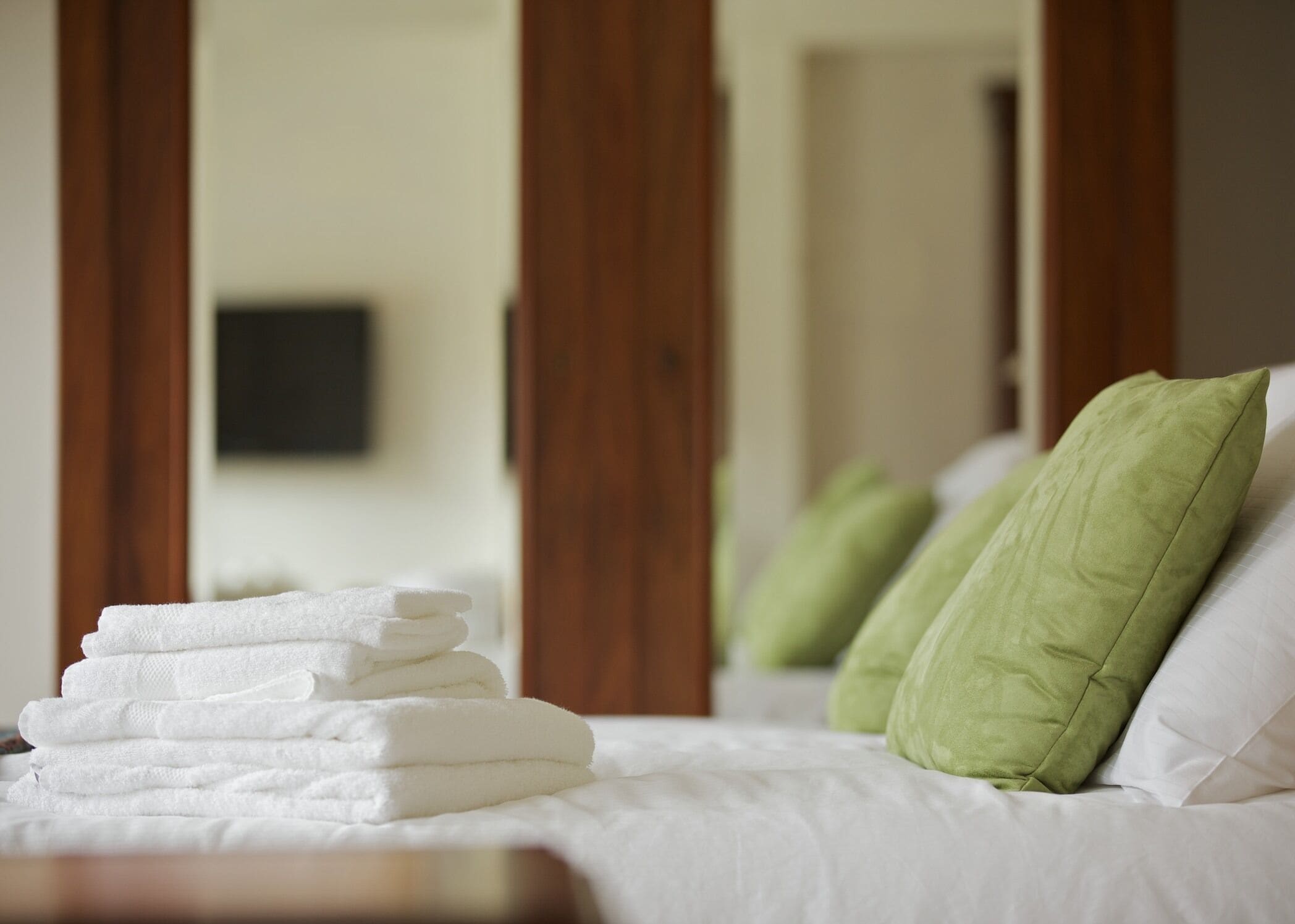 A neatly made bed with white linens and green pillows, a stack of folded towels, and wooden doors in the background.