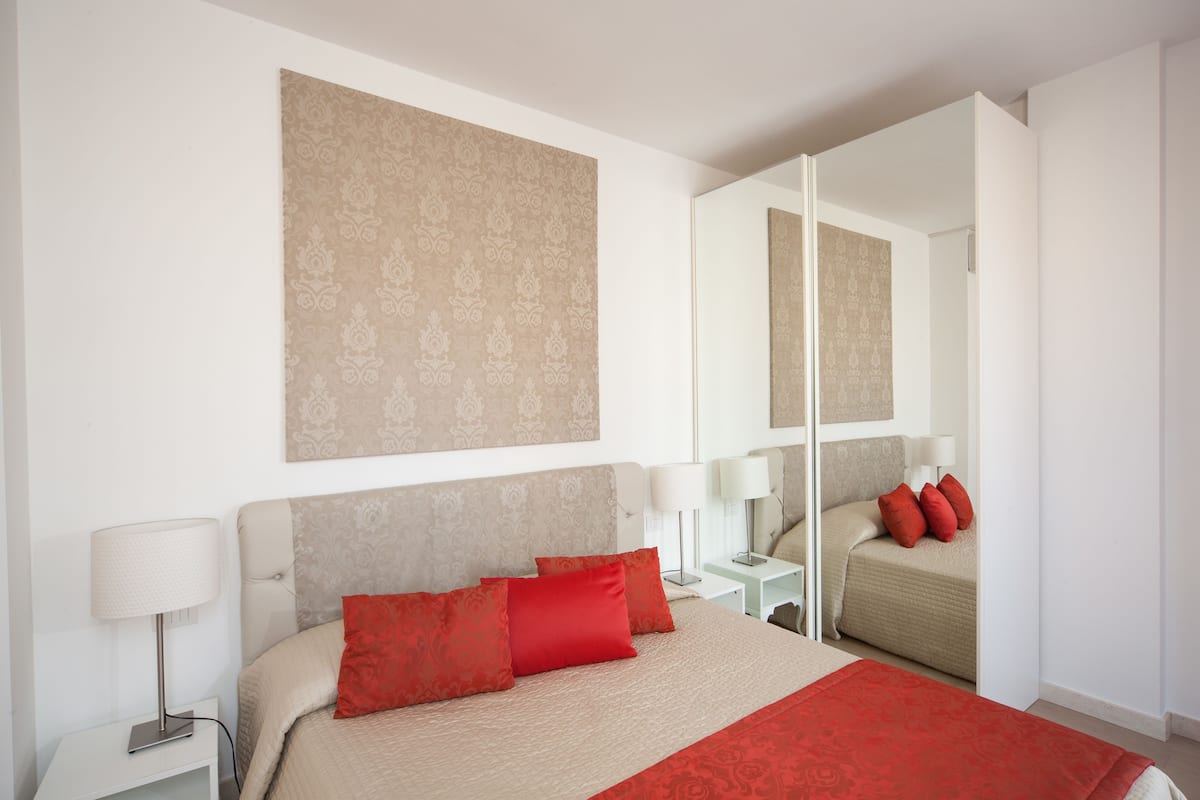 A modern hotel room with a large bed, a nightstand, a bedside table, and a mirror.