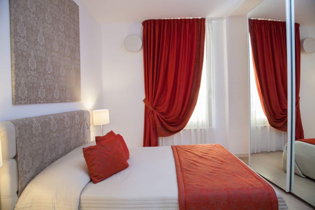 A hotel room with a bed, a bedside lamp, a mirror, and red curtains.