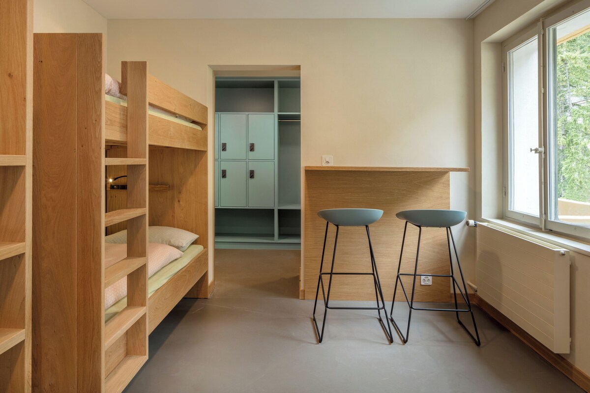 A room with bunk beds, a desk, two stools, and a window with a view.