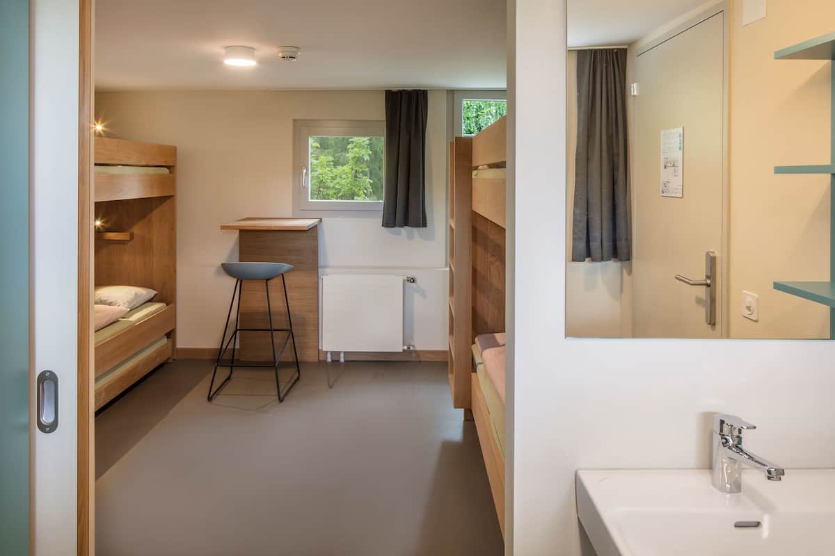 A compact room with bunk beds, a small stool, a window with curtains, a radiator, and a sink with a mirror.