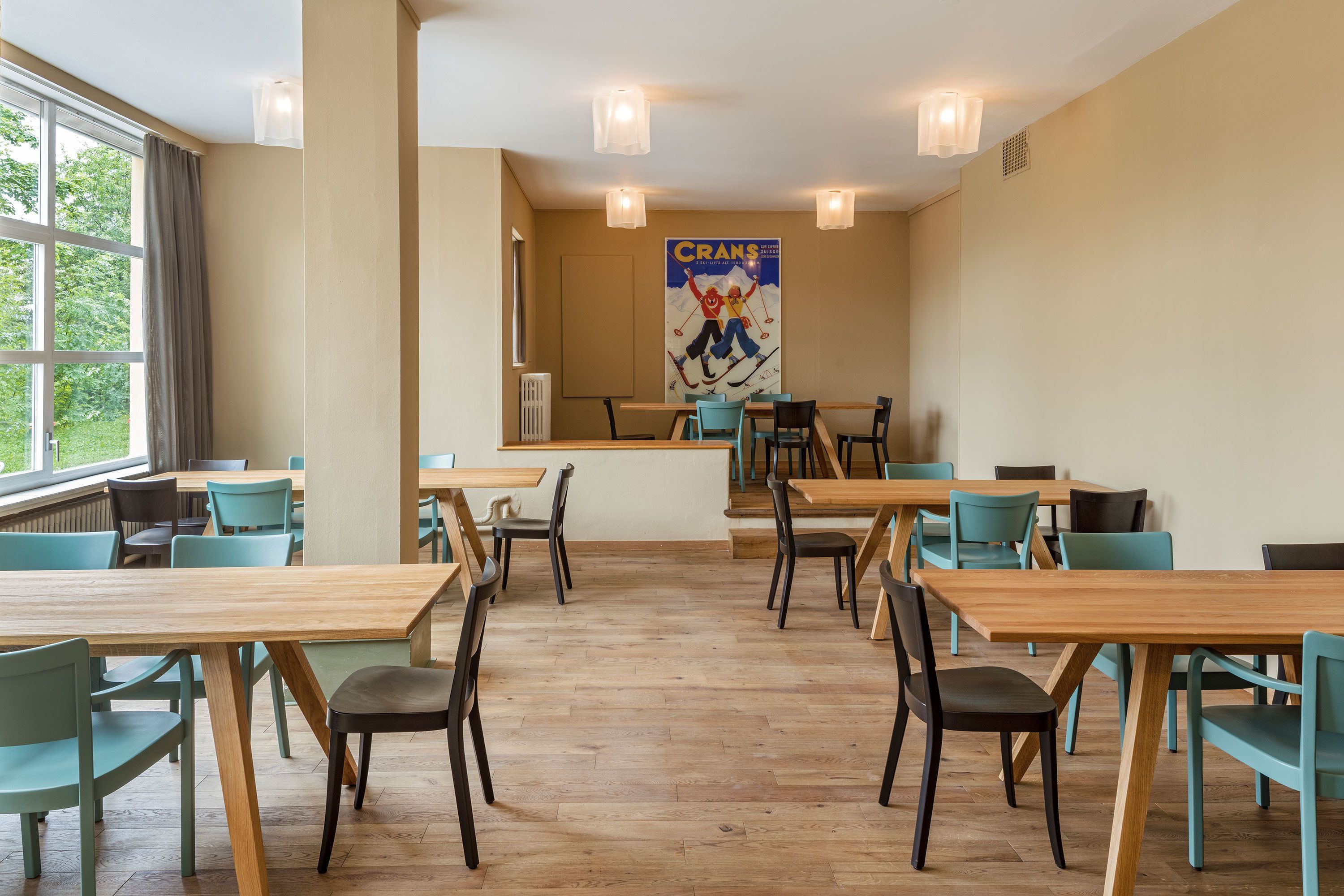 A spacious dining area with wooden tables and chairs, large windows, and a poster on the wall.