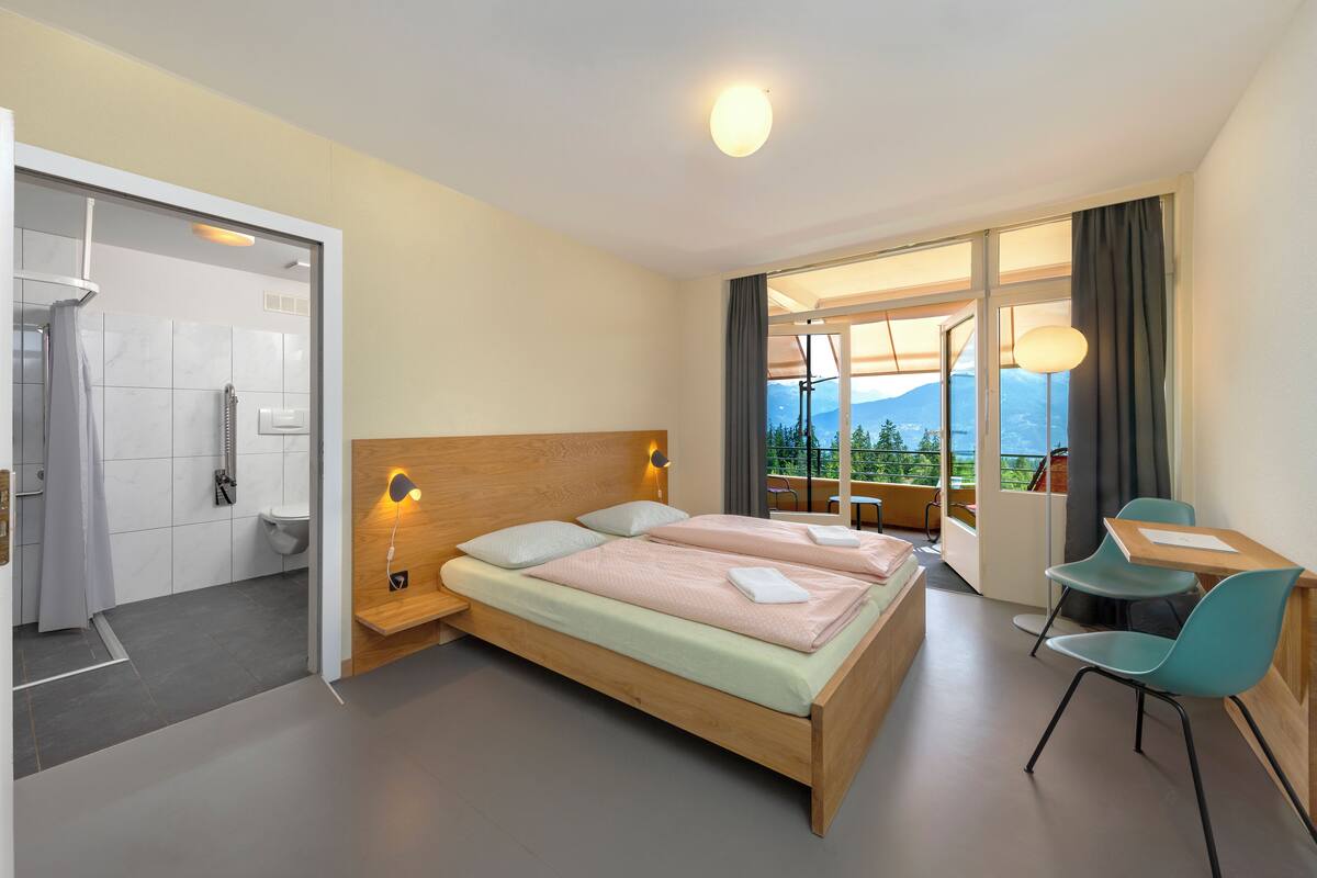 A modern hotel room with a wooden bed, a desk, and a view of mountains through the window.