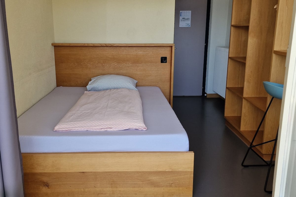A small, single-bed room with a wooden headboard, a grey mattress, and a light-colored blanket. There is a small wooden shelf unit to the right and a blue chair in the corner.