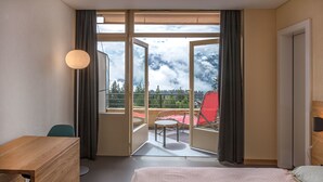 A hotel room with a bed, a desk, a chair, a table, and a balcony with a view of mountains. - Youth Hostel Crans-Montana (Crans-Montana)