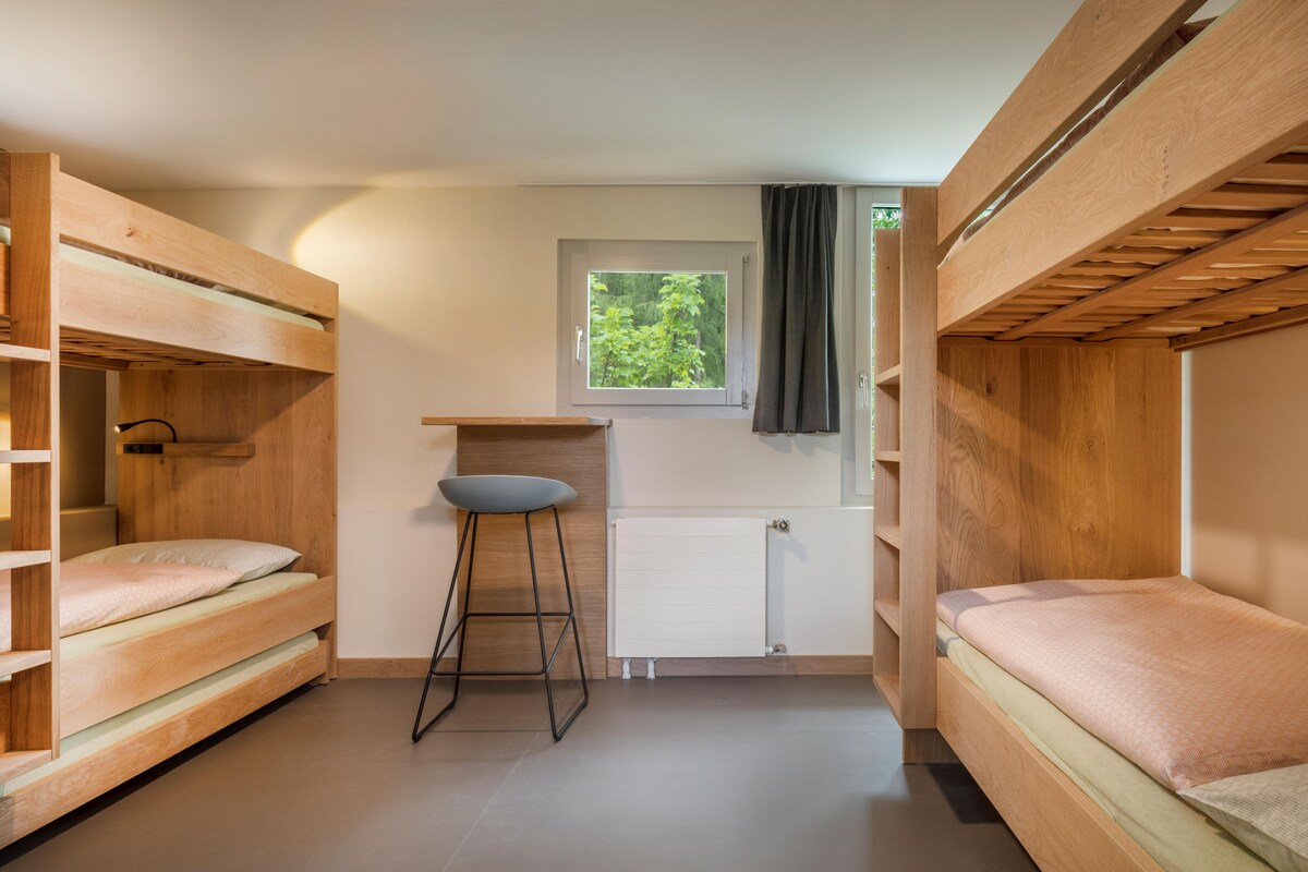 A compact room with two bunk beds, a small stool, a window with a view of greenery, and a radiator.